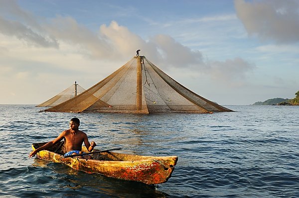 A young man in a canoe fishes near a fish farm at sunset.