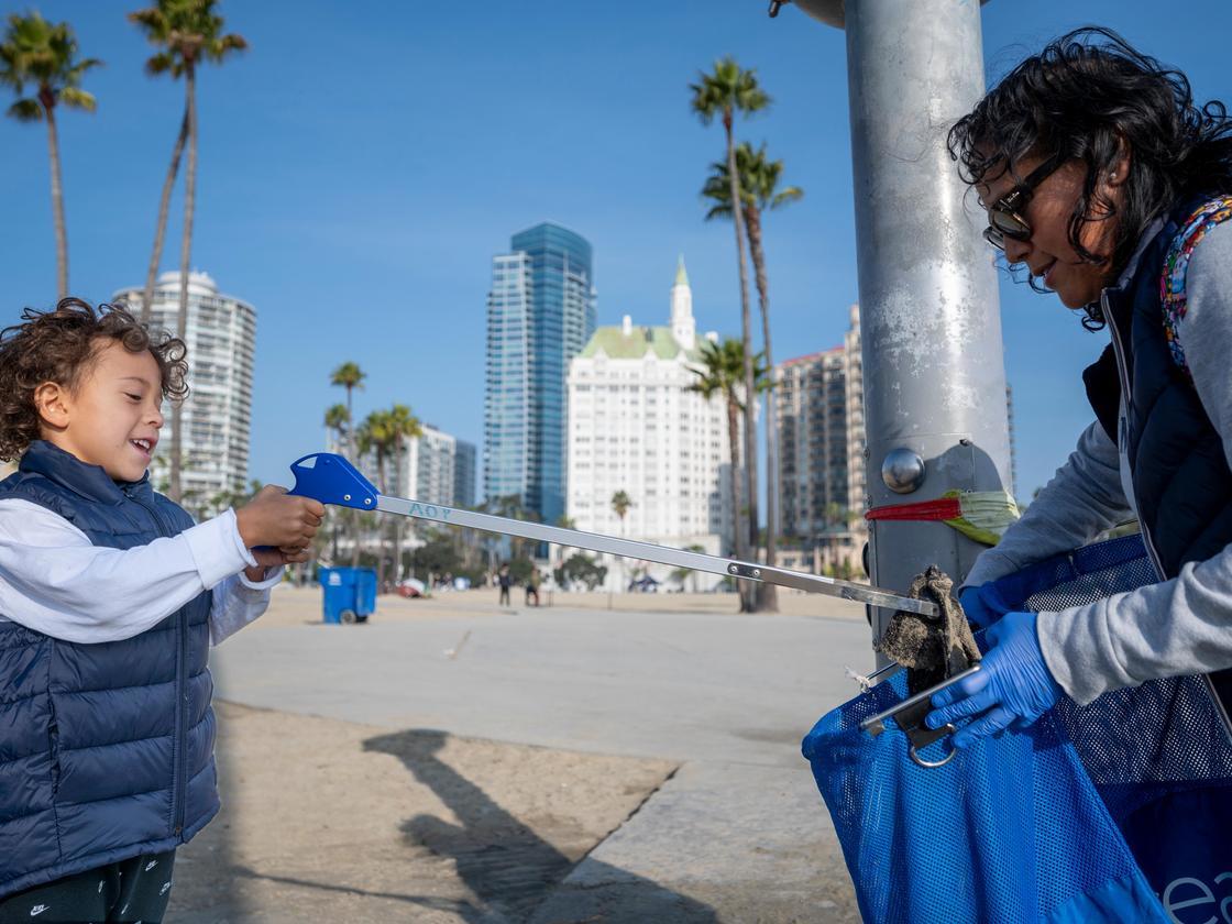 child wields a grabber to drop trash picked up off beach into a plastic bag behind held by an adult