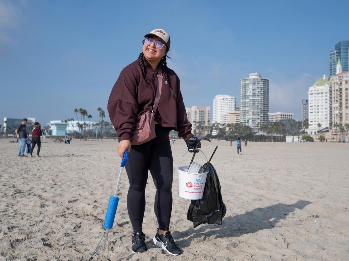 volunteer poses on a beach with a trash grabber in one hand and a bucket in the other