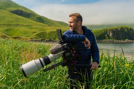 Ian Shive stands with a large nature photography camera on a tripod in tall grass in front of a lake and grassy hills