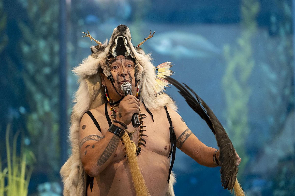 Chumash man in traditional indigenous attire, including a headdress with a skull, holding a microphone and a feathers, standing in front of a large aquarium.
