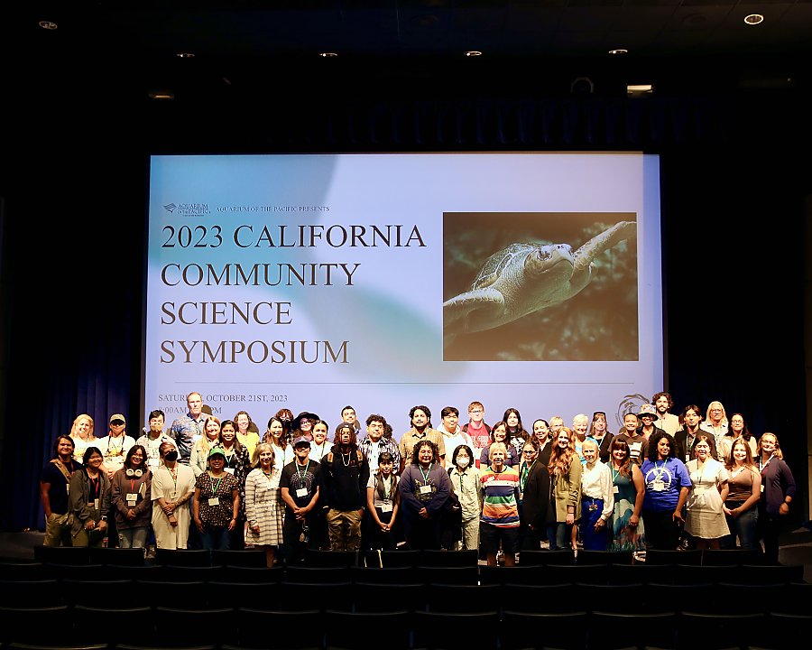 2023 CA Community Science Symposium on screen behind a group of dozens on a lit stage in a group photo