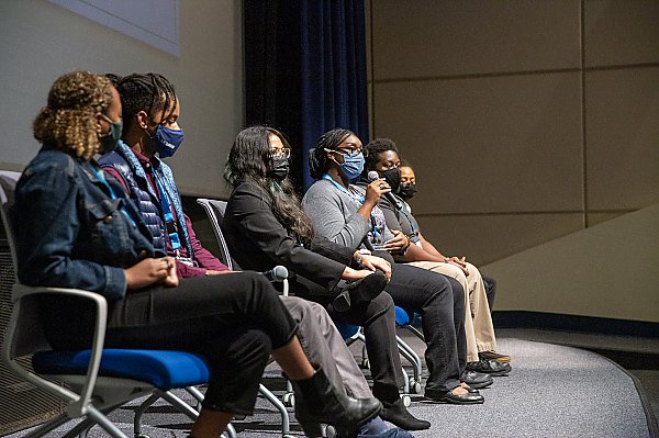 Six young black men and women sit on a stage with face masks on. One holds a mic as she talks to the audience just out of frame on the right.