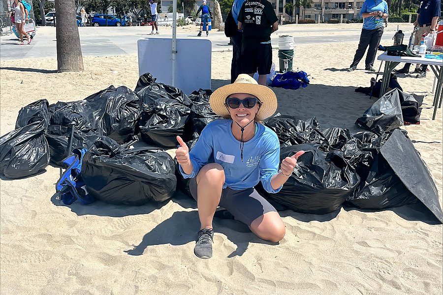 woman poses with thumbs up in front of a large pile of black garbage bags full of trash from a beach cleanup