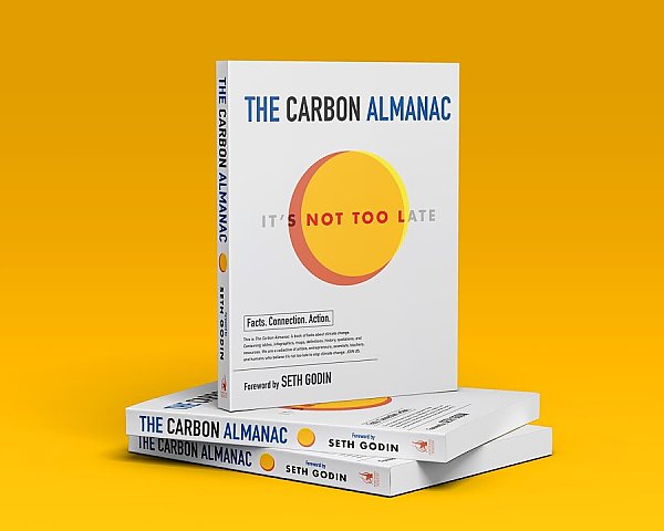 The Carbon Almanac book stands on top of two copies of the same book laid on the table