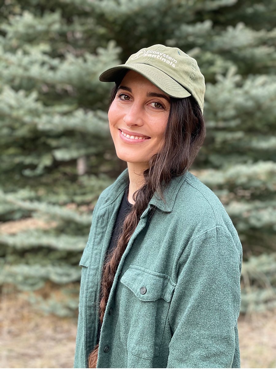 Photo of Caroline Cappello outdoors wearing a green cap and green flannel shirt over a black t-shirt.
