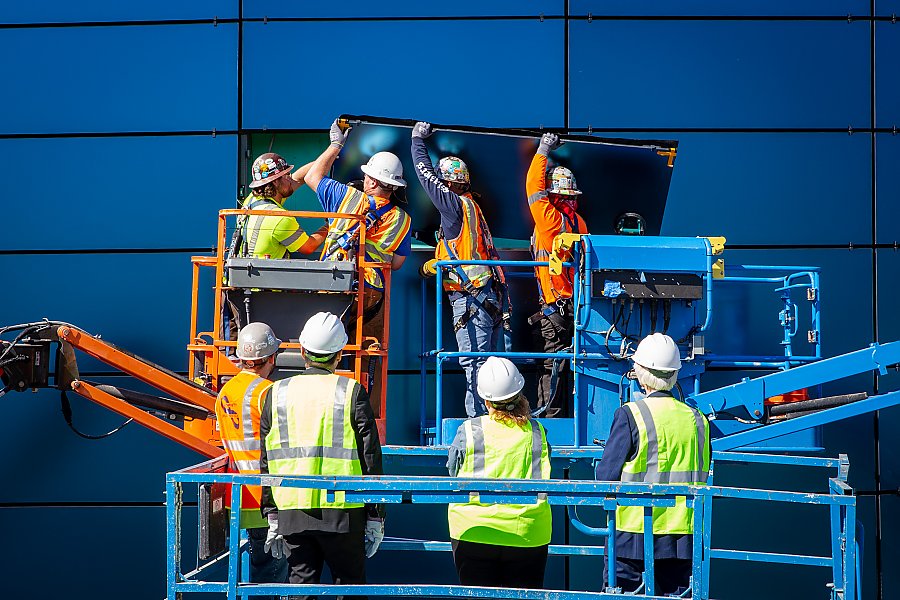 Crew on a lift installs glass panel in Pacific Visions facade while officials watch