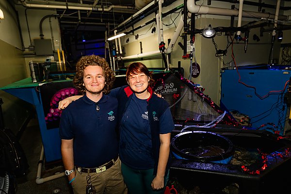 two aquarists standing side by side next to behind the scenes equipment