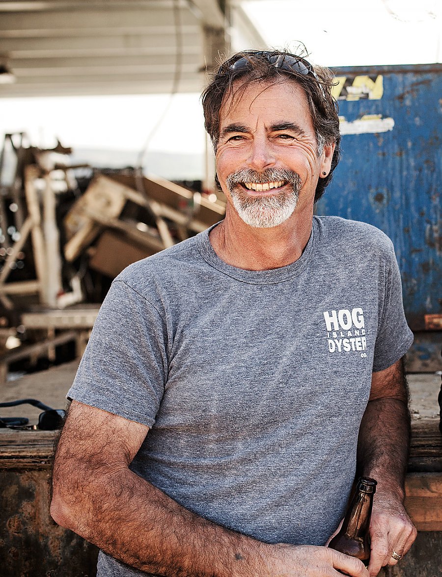 Hog Island Oyster farmer Terry Sawyer casually leans on a wood platform with farm equipment in the background. He is smiling and looking slightly to the right of the camera.
