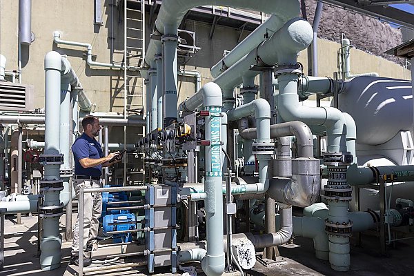 technician holds an electronic pad while inspecting pipes/plumbing in an outdoor industrial setting