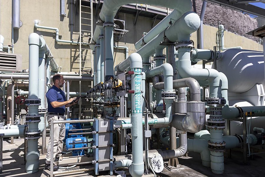technician holds an electronic pad while inspecting pipes/plumbing in an outdoor industrial setting