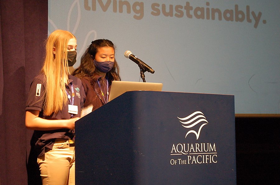 Two Five masked, uniformed, female, Teen Science Cafe volunteers give presentation at a podium in front of a large screen in a theater.
