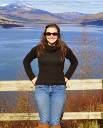Briana Warner with sunglasses, standing in front of a body of water with mountains in the background on a sunny fall day.